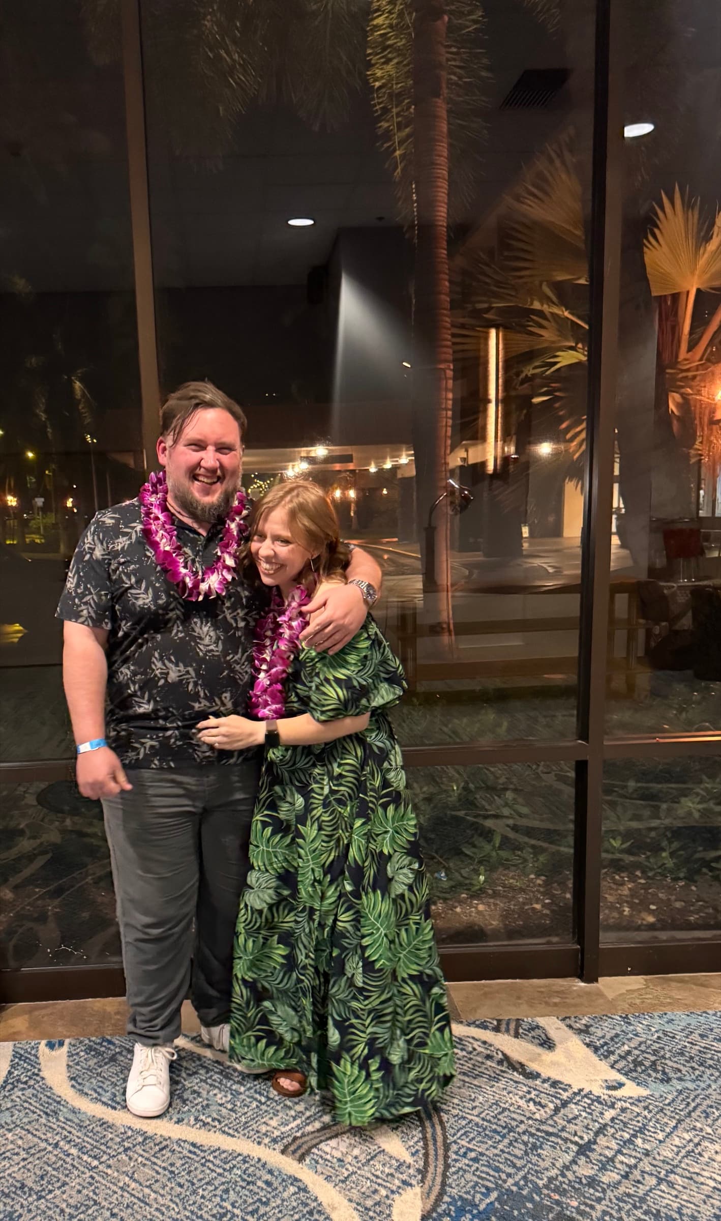 Kate and Matt wearing flower leis in front of windows at night.
