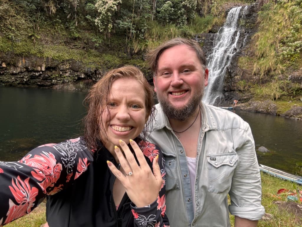 Kate and Matt beside a forest waterfall, with one holding up a ring.